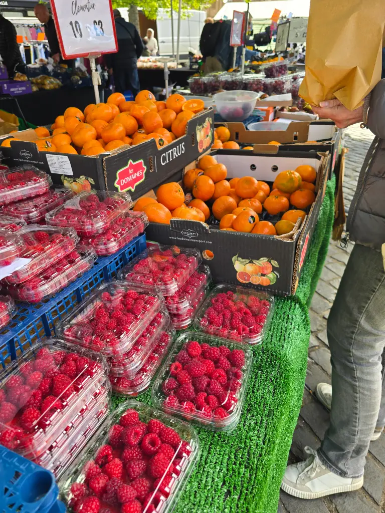 Outdoor market stall displaying boxes of oranges and stacked plastic punnets of raspberries on a green-covered table, with price signs above and a shopper holding a brown paper bag beside the stall